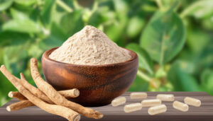 Powder supplement in a wooden bowl with dried root pieces and white capsules on a wooden surface.