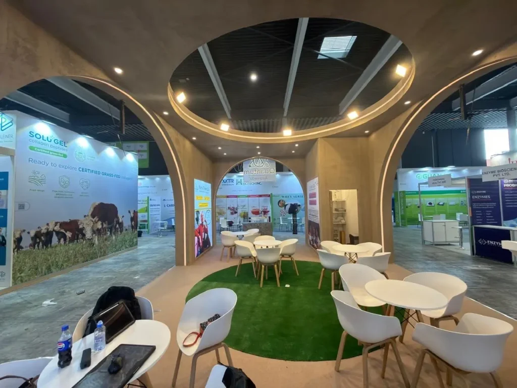 Exhibition booth with circular concrete arches, white tables and chairs around a green rug under circular ceiling rings.