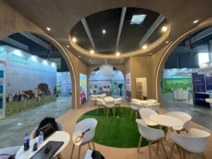 Exhibition booth with circular concrete arches, white tables and chairs around a green rug under circular ceiling rings.
