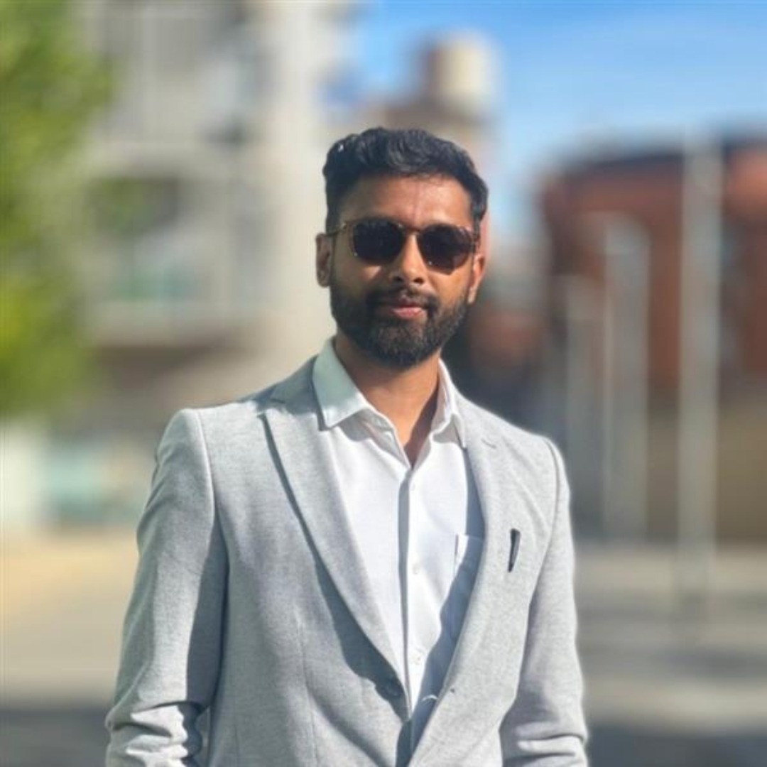 Man with a dark beard in a light gray blazer and white shirt, wearing sunglasses, standing outdoors with a blurred city background.