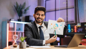Professional man in a suit presents a chart on a printed page, pointing with a green pen, at a tidy desk with a laptop and plants in a modern office.