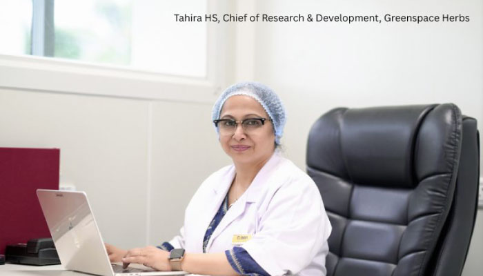 Tahira HS, Chief of Research & Development at Greenspace Herbs, sits at a desk wearing a lab coat and hairnet with a laptop.
