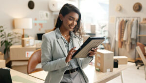 Businesswoman in a light gray blazer uses a tablet in a bright shop workspace with boxes and clothing racks in the background.