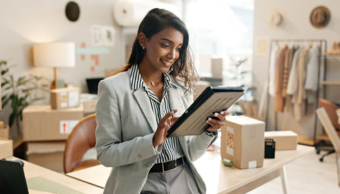 Businesswoman in a light gray blazer uses a tablet in a bright shop workspace with boxes and clothing racks in the background.