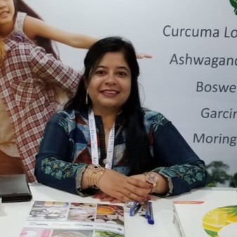 Smiling woman seated at a booth with brochures and pens on the table, wearing a lanyard, with a display behind her.