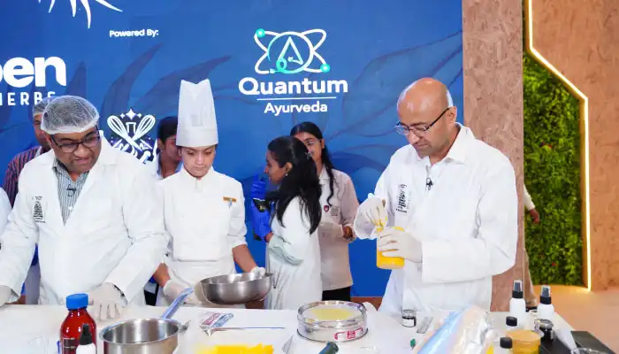 Chefs and volunteers in white coats conduct a cooking demo at a Quantum Ayurveda booth, with pots, bottles, and a blue event backdrop displaying the logo.