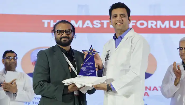 Two men on stage during an award ceremony; a man in a white lab coat hands a star-shaped trophy to a man in a dark suit, with a banner and audience in the background.
