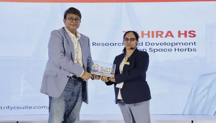 Two professionals on stage shake hands as a man hands a plaque to a woman during an awards ceremony, with lanyards visible.