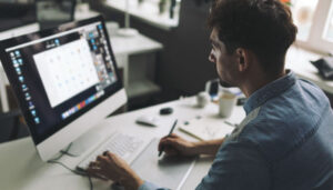 Man in a blue shirt sits at a desk, working on a desktop computer with a large monitor in a bright office.