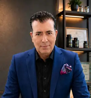 Man in a blue blazer and black shirt, posing for the camera in a modern office setting with shelves in the background.
