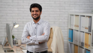 Smiling man in a striped shirt stands with arms crossed in a modern office, with a desk and shelves in the background.
