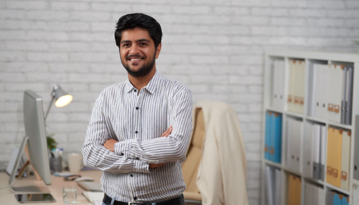 Smiling man in a striped shirt stands with arms crossed in a modern office, with a desk and shelves in the background.