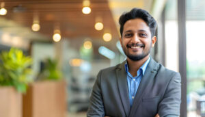 Smiling man in a gray suit and blue shirt stands in a bright open-plan office with warm lighting behind him.
