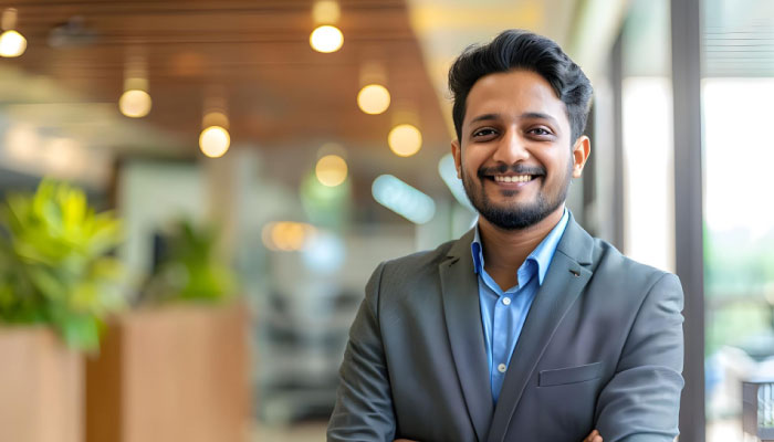 Smiling man in a gray suit and blue shirt stands in a bright open-plan office with warm lighting behind him.